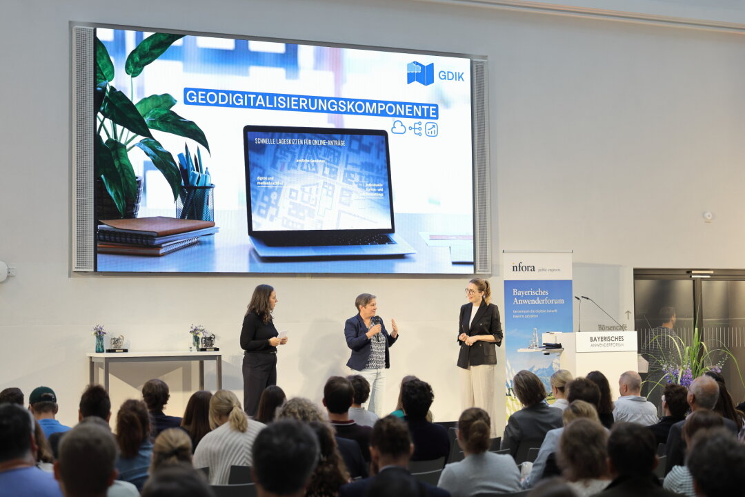 Konferenzsaal mit drei Frauen auf der Bühne unter einer großen Leinwand, auf der unter dem Titel 